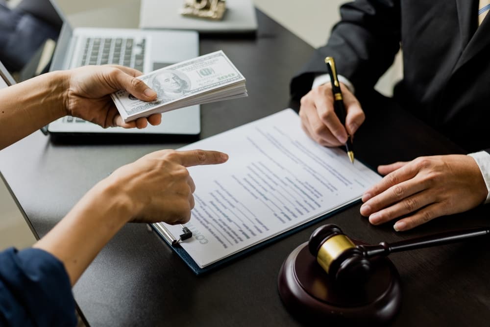 Business people shaking hands discreetly exchange money with an attorney under the table, attempting to influence a court case outcome.