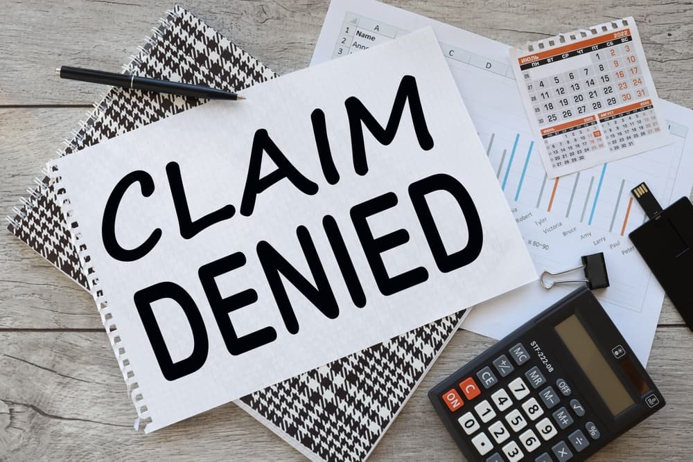 A work surface displays a piece of paper with the bold text "CLAIM DENIED," next to a calculator and a calendar. 