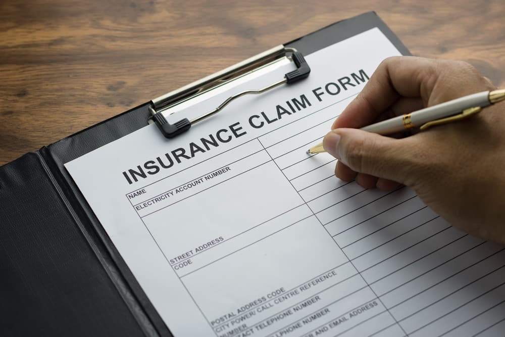 A hand holding a pen is filling out an application form for registering an insurance claim.