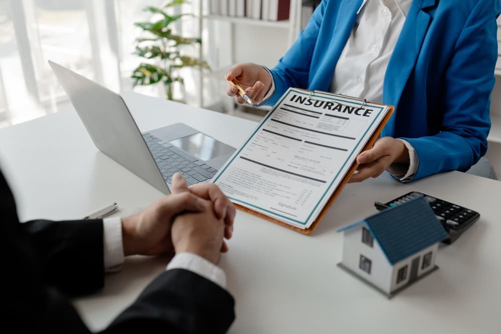 A realtor is seated in their office, explaining home insurance documents to a buyer. The paperwork is spread out on the desk, including a house insurance policy and various forms.