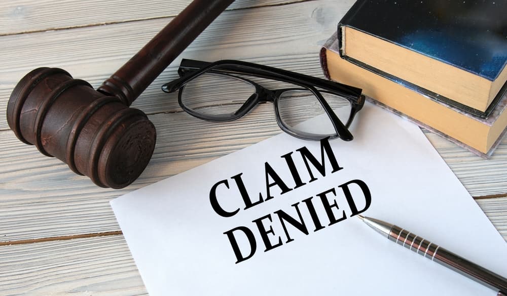 "CLAIM DENIED" written on white paper, set against a background featuring a judge's gavel, eyeglasses, and a pen.