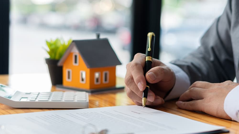 Real estate office agents handle contract documents alongside house designs and calculators on the table, assisting clients with home sales.